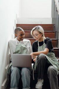 Young smiling diverse female friends in aprons sitting on stairs and working on project using laptop